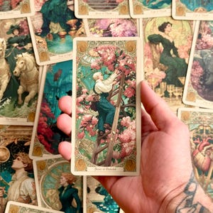 Op de afbeelding: Een rechthoekige tarotkaart getiteld Scorn of Pleiades met een illustratie van een persoon in een donkergroene tuinbroek die een houten ladder beklimt om roze bloemen in een kas te bereiken, omlijst door een sierlijke gouden rand.