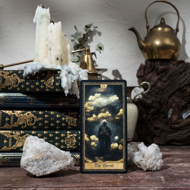 May include: A tarot card reading featuring the The Hermit card from a tarot deck. The card is black with gold accents and features a dark figure in a hooded robe standing in a landscape of clouds. The card is sitting on a wooden table with a stack of books, a candle, and a teapot in the background.