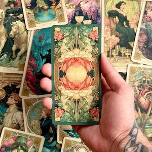 Op de afbeelding: Een rechthoekige tarotkaart met een symmetrisch, sierlijk ontwerp met een centrale gouden cirkel omringd door gelaagde, gestileerde koraalroze en cr&egrave;mekleurige bloemmotieven, alles tegen een diepgroene achtergrond met ingewikkeld goud filigraan.
