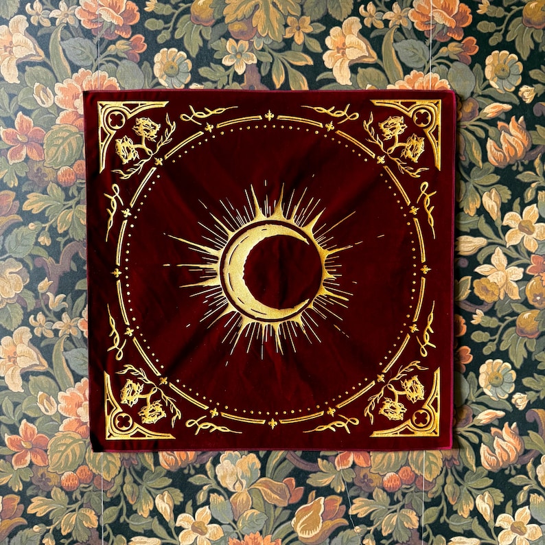 May include: A red velvet bandana with a gold sun and crescent moon design. The bandana has a decorative border with a repeating pattern.