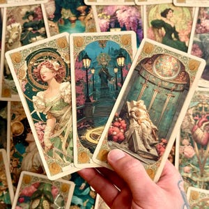 Op de afbeelding: Drie tarotkaarten met ingewikkelde art-nouveau-randen. De linkerkaart toont een geblinddoekte figuur in een groene jurk die een zwaard vasthoudt. De middelste kaart toont een standbeeld in een park met lantaarnpalen. De rechterkaart toont een figuur die rust op een stenen plaat onder drie zwaarden.