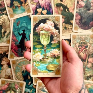 Op de afbeelding: Een rechthoekige tarotkaart getiteld Ace of Cups met een centrale versierde gouden kelk gevuld met gloeiende vloeistof, omringd door witte waterlelies en groene bladeren op een blauwgroene vijver, omlijst door een decoratieve rand met gouden accenten.