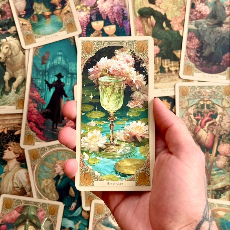 Op de afbeelding: Een rechthoekige tarotkaart getiteld Ace of Cups met een centrale versierde gouden kelk gevuld met gloeiende vloeistof, omringd door witte waterlelies en groene bladeren op een blauwgroene vijver, omlijst door een decoratieve rand met gouden accenten.