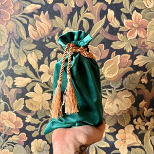 May include: A green velvet drawstring bag with gold rope and tassels. The bag is held in a hand.