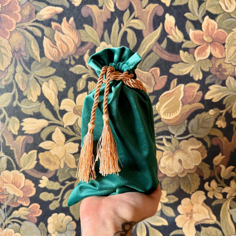 May include: A green velvet drawstring bag with gold rope and tassels. The bag is held in a hand.