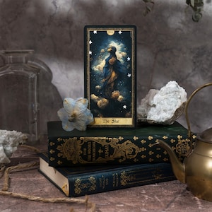 May include: A tarot card with a dark blue background and gold accents. The card depicts a woman standing in a pool of water, with stars in the sky above her. The card is titled "The Star" and is numbered XVII. The card is sitting on top of a stack of books.