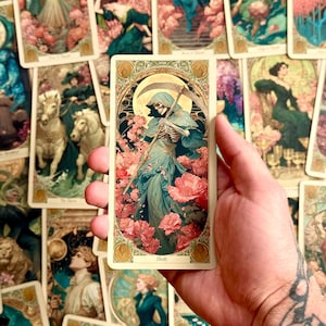 Op de afbeelding: Een rechthoekige tarotkaart met een illustratie van een skeletfiguur in een blauwe mantel, die een zeis vasthoudt te midden van roze bloemen, met het woord Death onderaan, binnen een sierlijke goudkleurige rand.