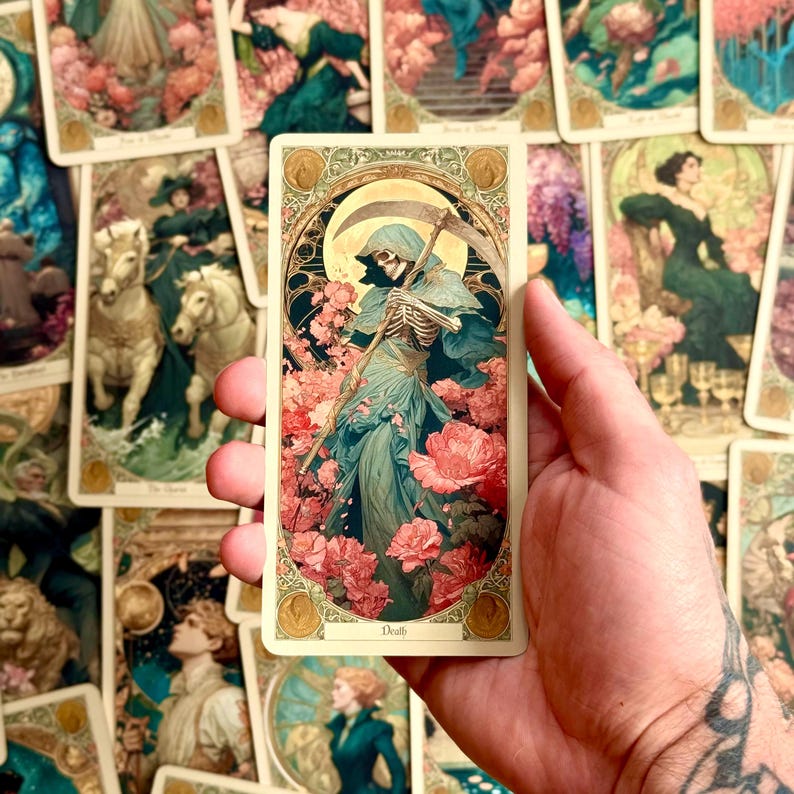 Op de afbeelding: Een rechthoekige tarotkaart met een illustratie van een skeletfiguur in een blauwe mantel, die een zeis vasthoudt te midden van roze bloemen, met het woord Death onderaan, binnen een sierlijke goudkleurige rand.