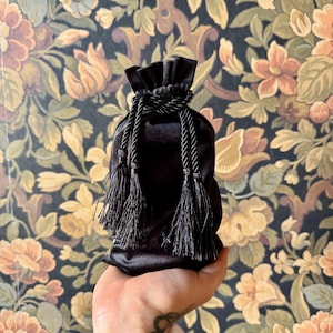May include: A black velvet drawstring bag with black tassels. The bag is held in a hand.