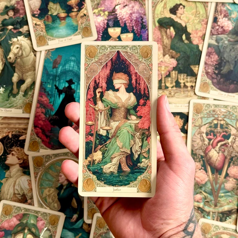 Op de afbeelding: Een rechthoekige tarotkaart met de titel Justice, voorzien van een illustratie van een geblinddoekte figuur in een groene jurk die een gouden weegschaal vasthoudt, omringd door roze bloemaccenten en een sierlijke rand, tegen een achtergrond van andere tarotkaarten.