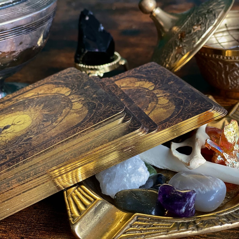May include: A stack of gold-edged tarot cards with intricate designs, resting on a brass dish filled with various crystals and a bone-like object.