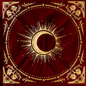 May include: A red velvet bandana with a gold crescent moon and sun design. The bandana has a decorative border with a repeating pattern of leaves and flowers.