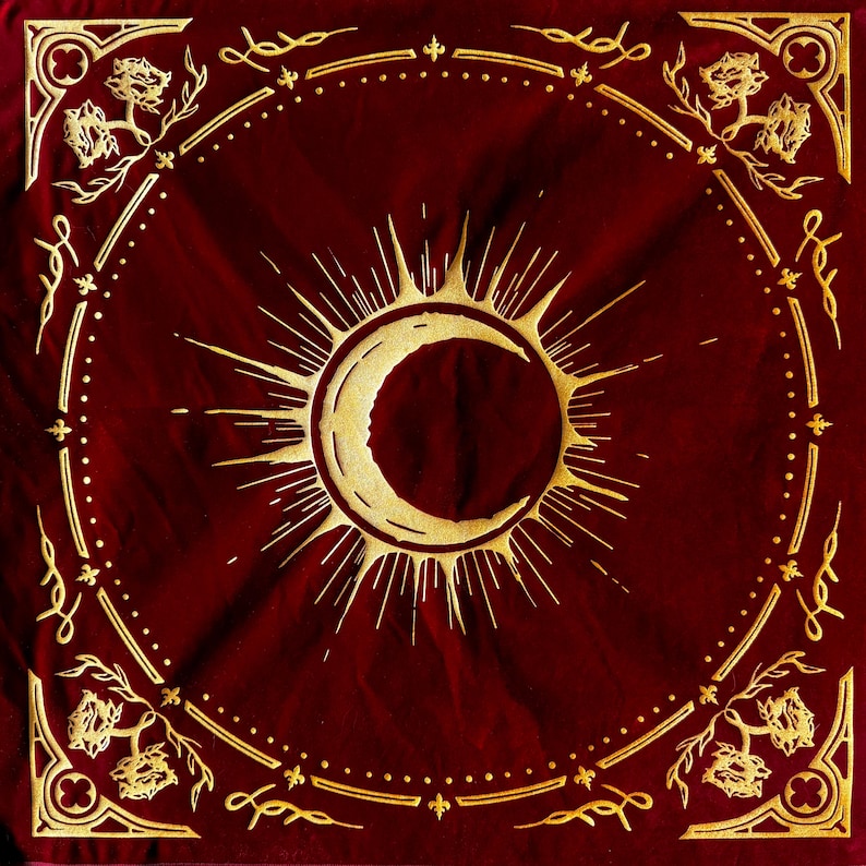 May include: A red velvet bandana with a gold crescent moon and sun design. The bandana has a decorative border with a repeating pattern of leaves and flowers.