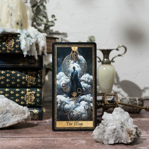 May include: A tarot card with a black background and gold accents. The card depicts a figure in a dark robe standing on a cloud with a staff in their hand. The card is titled "The Mage".