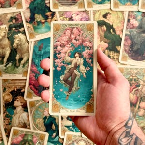 Op de afbeelding: Een rechthoekige tarotkaart getiteld The Hanged Man met een illustratie van een persoon die aan &eacute;&eacute;n been aan een tak hangt te midden van roze kersenbloesems boven blauwgroen water, omlijst door een sierlijke gouden rand.