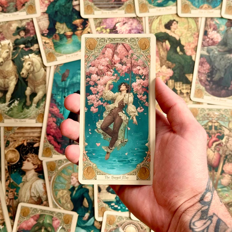 Op de afbeelding: Een rechthoekige tarotkaart getiteld The Hanged Man met een illustratie van een persoon die aan &eacute;&eacute;n been aan een tak hangt te midden van roze kersenbloesems boven blauwgroen water, omlijst door een sierlijke gouden rand.