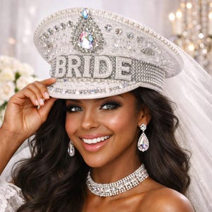 Puede incluir: Un sombrero de novia blanco con la palabra "BRIDE" en letras plateadas, adornado con pedrería y cristales. La novia lleva un collar y pendientes de diamantes. Un velo está sujeto al sombrero.