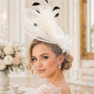 May include: A white headpiece with a veil, feathers, and a flower. The headpiece features a delicate net veil that drapes over the face. The design includes white feathers and a white flower, with accents of black feathers.