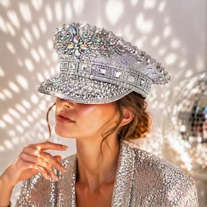 Pode incluir: Um chapéu prateado adornado com strass e cristais brilhantes. O chapéu apresenta uma aba e uma coroa estruturadas, cobertas por detalhes brilhantes. O chapéu faz parte de uma coleção de acessórios glamourosos.