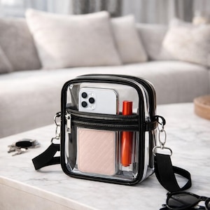May include: A clear crossbody bag with a black trim and strap. The bag contains a smartphone, a pink cardholder, and a tube of orange lipstick. The bag is sitting on a white marble surface.