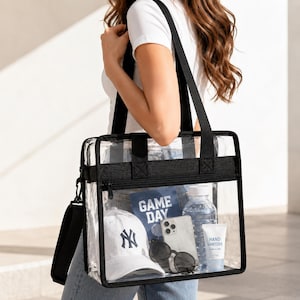 May include: A clear tote bag with black trim, carrying a white baseball cap with a navy blue logo, sunglasses, a smartphone, a water bottle, and hand sanitizer. The bag also contains a blue booklet that says "GAME DAY".