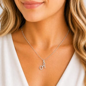 May include: A silver necklace featuring a pendant shaped like the letter "A", adorned with small, sparkling stones. The necklace is worn around the neck, with the pendant resting on the chest.