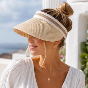 May include: A beige straw visor featuring a wide, structured brim and a thick, woven headband. The headband is accented with a horizontal stripe pattern in shades of white and light brown, providing a clean, neutral aesthetic for sun protection.