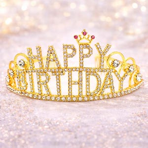 May include: A gold-colored tiara with the words "HAPPY BIRTHDAY" spelled out in clear, sparkling rhinestones. The crown features a small crown design at the top with pink gems. The tiara is designed for a birthday celebration.