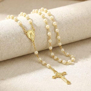 Puede incluir: Un collar de rosario dorado con cuentas de perlas de imitación. El collar presenta un colgante de crucifijo y una pieza central con una figura religiosa. Las cuentas están ensartadas en una cadena dorada.