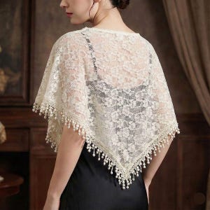 Op de afbeelding: Ivoor kanten sjaal met een delicaat bloemmotief en een geschulpte rand. De sjaal is afgezet met kleine, bungelende kralen. Het kledingstuk wordt gedragen over een zwarte top, wat een contrast in kleur en textuur cre&euml;ert. Een formeel accessoire.