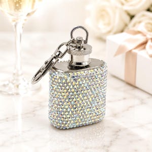 May include: A rectangular, silver-toned flask covered in iridescent rhinestones. The flask has a screw-on cap, a small loop, and a hinged clasp. The background includes a glass of champagne and a gift box.