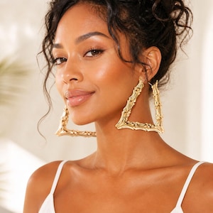 May include: Gold-colored bamboo-style triangle hoop earrings. The earrings are large and have a textured surface, reflecting light. The earrings are worn on a person.