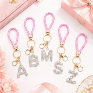Puede incluir: Llaveros de cuero sintético rosa con herrajes dorados y letras incrustadas de diamantes de imitación. Las letras son A, B, M, S y Z. Los llaveros se muestran sobre una superficie blanca con accesorios rosas.