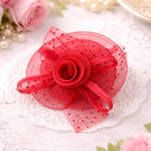 May include: A vibrant red hair accessory featuring a central rose and a layered design. The accessory includes a sheer, dotted fabric and ribbon accents, resting on a white lace doily. This elegant piece is suitable for special occasions.