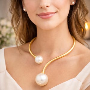 May include: A gold-toned necklace with two large, white, round pearls. The necklace has a curved design that sits around the neck. A matching pearl earring is visible on the left side of the image. The background is blurred.