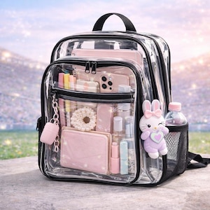 Puede incluir: Una mochila transparente con ribetes negros, llena de accesorios rosas, incluyendo un teléfono, una cartera y cosméticos. Un charm de conejito rosa y una botella de agua están adjuntos. La mochila tiene un asa superior y un bolsillo lateral de malla.