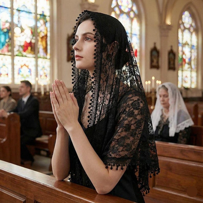 Op de afbeelding: Een zwarte kanten sluier met een delicaat bloemmotief en een kralenrand. De sluier is over het hoofd en de schouders gedrapeerd. De foto is genomen in een kerk met glas-in-loodramen.