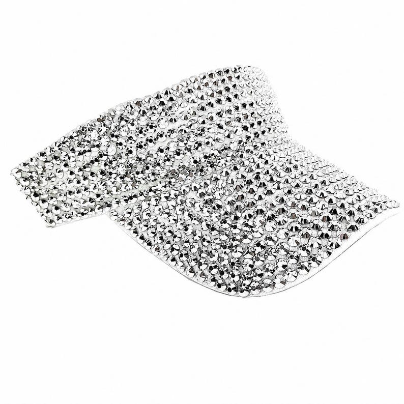 Puede incluir: Una visera plateada cubierta de peque&ntilde;os y brillantes pedrer&iacute;a. La visera es curvada y tiene una ala ancha para dar sombra. La pedrer&iacute;a refleja la luz, creando un efecto deslumbrante. Este accesorio est&aacute; dise&ntilde;ado para ser usado en la cabeza.