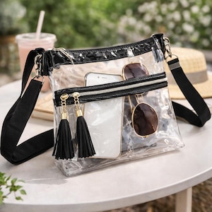May include: Transparent crossbody bag with a black trim and strap. The bag has a zipped pocket with gold-coloured zipper pulls and black tassels. Inside the bag are a white smartphone and sunglasses.