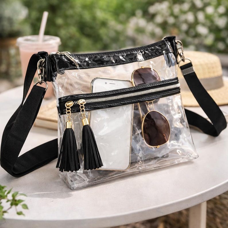 May include: Transparent crossbody bag with a black trim and strap. The bag has a zipped pocket with gold-coloured zipper pulls and black tassels. Inside the bag are a white smartphone and sunglasses.