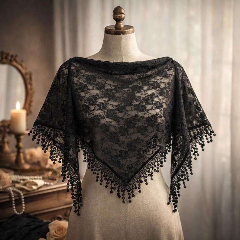 Op de afbeelding: Een zwarte kanten sjaal met een bloemmotief, gepresenteerd op een mannequin. De sjaal heeft een vierkante vorm en is afgewerkt met een kralenfranje. De achtergrond bevat een spiegel, een brandende kaars en een houten dressoir.