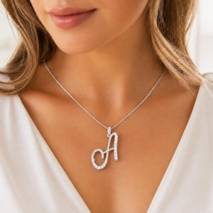 Puede incluir: Un collar plateado con un colgante con la forma de la letra "A". El colgante está incrustado con pequeñas piedras brillantes. El collar se lleva alrededor del cuello.