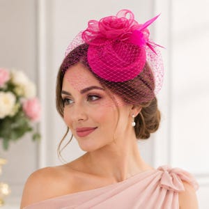 Op de afbeelding: Een felroze fascinator met een rozenaccent, net en een veer. De hoofddeksel wordt gedragen door een persoon in een roze jurk. Een formele hoofddeksel voor speciale gelegenheden.