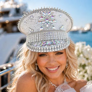May include: A silver sequined captain's hat with crystal and rhinestone embellishments. The hat features a flat top, a wide brim, and a decorative band. The hat is adorned with sparkling crystals and rhinestones, creating a glamorous and eye-catching accessory.