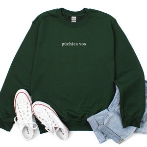 May include: A dark green sweatshirt with the text "p&uacute;chica vos" in white lettering. The sweatshirt is laid flat on a white background with a pair of white sneakers and blue jeans.