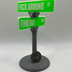 以下が含まれることがあります： 緑色の背景に白い文字で「FUCK AROUND ST」と「FIND OUT BLVD」と書かれた3Dプリントされた街路標識。標識は灰色のポールに灰色の土台で取り付けられています。