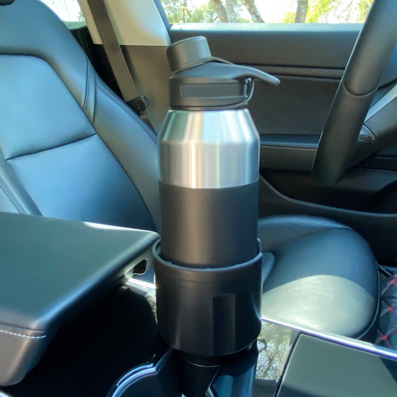Op de afbeelding: Een roestvrijstalen waterfles met een zwarte dop en een zwarte siliconen houder. De fles staat in een bekerhouder in de auto.