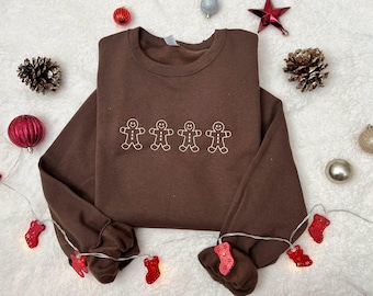 Pode incluir: Um moletom marrom com quatro bonecos de gengibre bordados. A imagem é decorada com enfeites de Natal, incluindo enfeites, pinhas e uma corda de luzes com lâmpadas vermelhas em forma de meia.