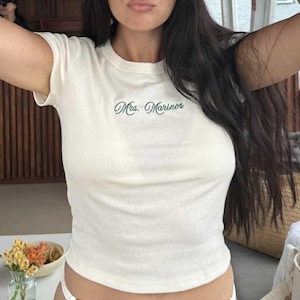 May include: Cream-colored, short-sleeved t-shirt with the text "Mrs. Marinos" in green script. The shirt has a crew neck and a fitted silhouette. The image shows a close-up of the shirt.