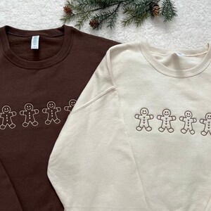 Może przedstawiać: Dwie bluzy z długimi rękawami z haftem piernikowego ludzika. Jedna jest brązowa, a druga kremowa.